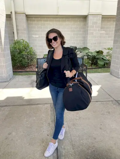 Sherry com bolsa e mochila no aeroporto com jaqueta preta e blusa preta e jeans