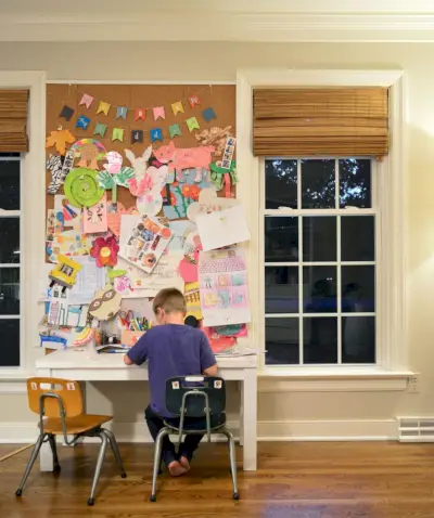 Hijo trabajando en un pequeño espacio de escritorio de arte en sillas preescolares con un gran espacio de exhibición de corcho entre las ventanas