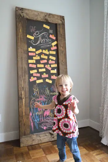 Niña pequeña parada frente a una pizarra inclinada de bricolaje con notas de gratitud de Acción de Gracias