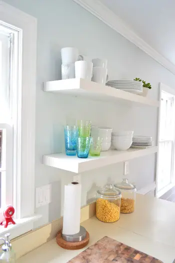 Dos largos estantes flotantes de Ikea en una cocina con tazas coloridas y pilas de vajilla blanca.