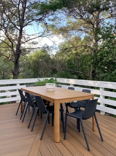 Mesa de comedor al aire libre de metal y madera sintética con sillas negras rodeadas de árboles