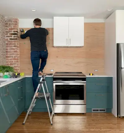 John em pé na escada pregando backsplash com painéis de madeira na cozinha azul