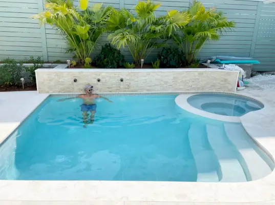 John em uma pequena piscina de forma livre contra um muro de contenção com detalhes em pedra sob palmeiras