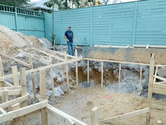 Agujero de piscina enmarcado con madera y John al fondo con cinta métrica