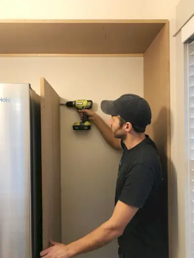 John perce un panneau MDF vertical dans du bois déjà placé près du réfrigérateur dans le garde-manger