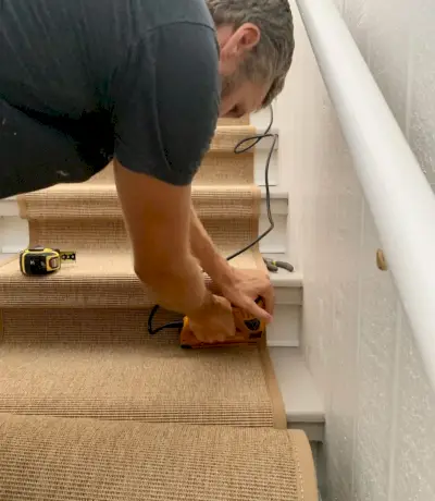 John utilise une agrafeuse électrique pour enfoncer l'agrafe dans le coin inférieur de l'escalier afin de sécuriser le tapis en sisal