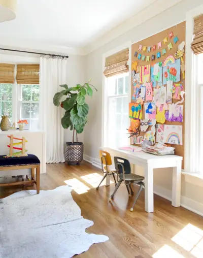 Espaço de escritório com mesa infantil com janelas e painel de cortiça do tamanho de uma parede acima da mesa infantil