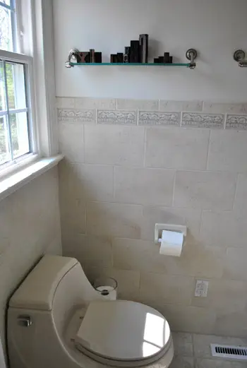 Vue sur mur carrelé dans une salle de bains datée avec étagère en verre au-dessus des toilettes